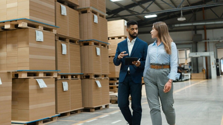 Two supply chain professionals walking through a warehouse with stacked cardboard pallets, discussing logistics operations and inventory strategy