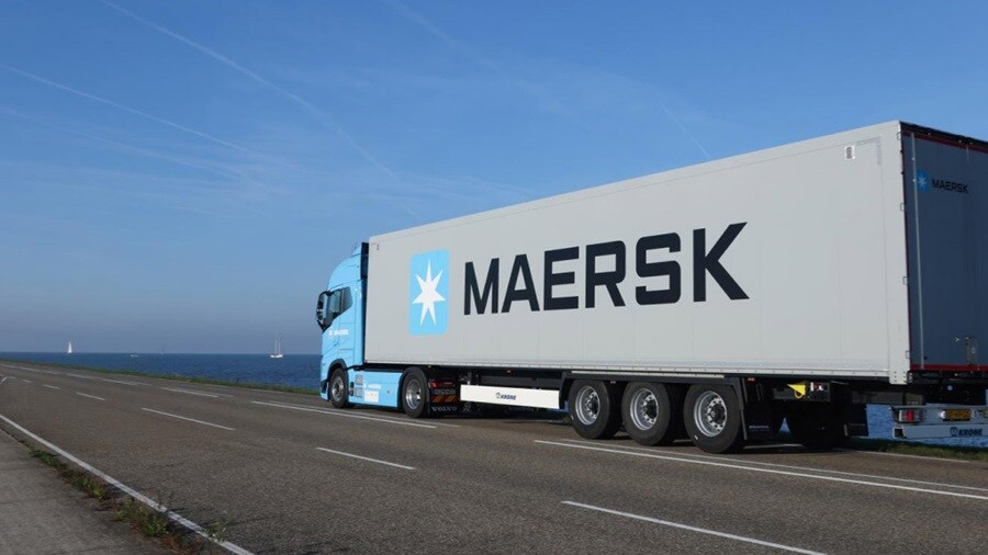 A Maersk-branded truck drives along a coastal road under a clear blue sky.