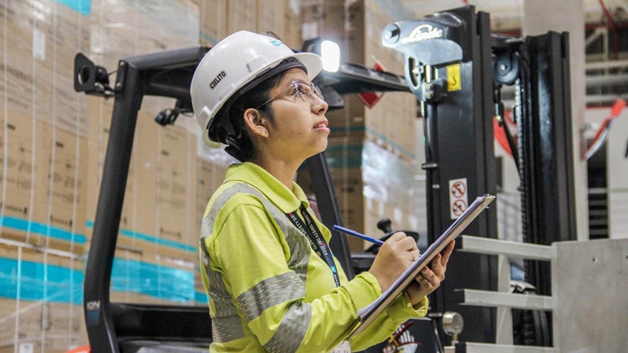 A woman in a safety vest stands beside a forklift in a warehouse setting.