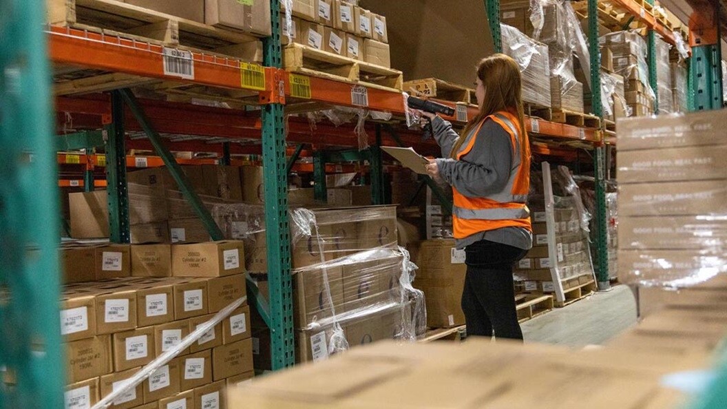 A woman wearing an orange vest stands in a warehouse, surrounded by shelves and boxes.