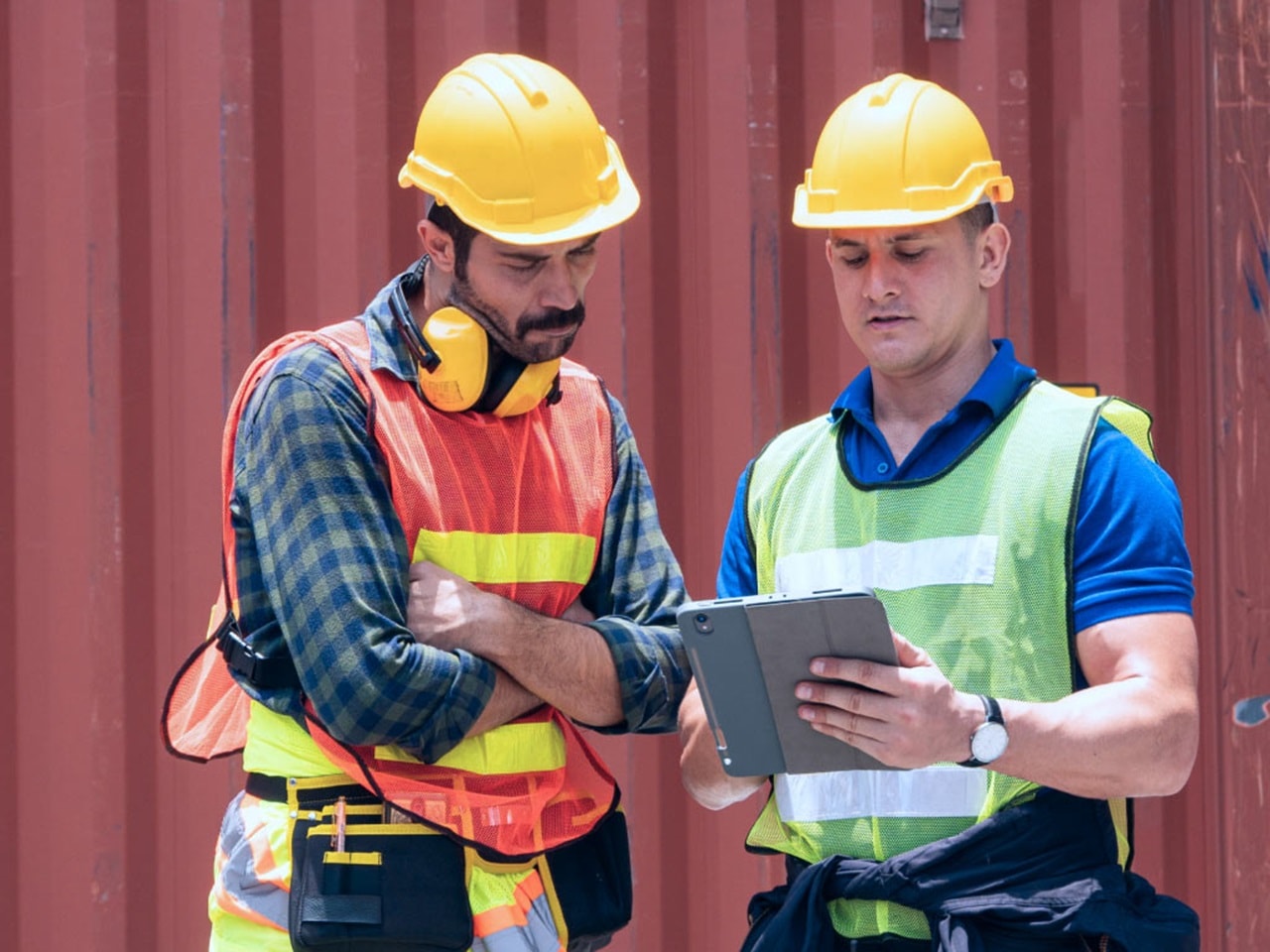Two workers wearing safety helmets and vests reviewing supply chain operations on a digital tablet at container yard