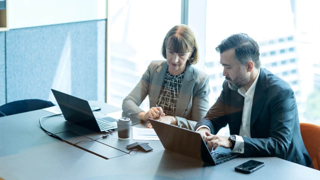 Two business professionals collaborating on laptops in office meeting to discuss supply chain solutions